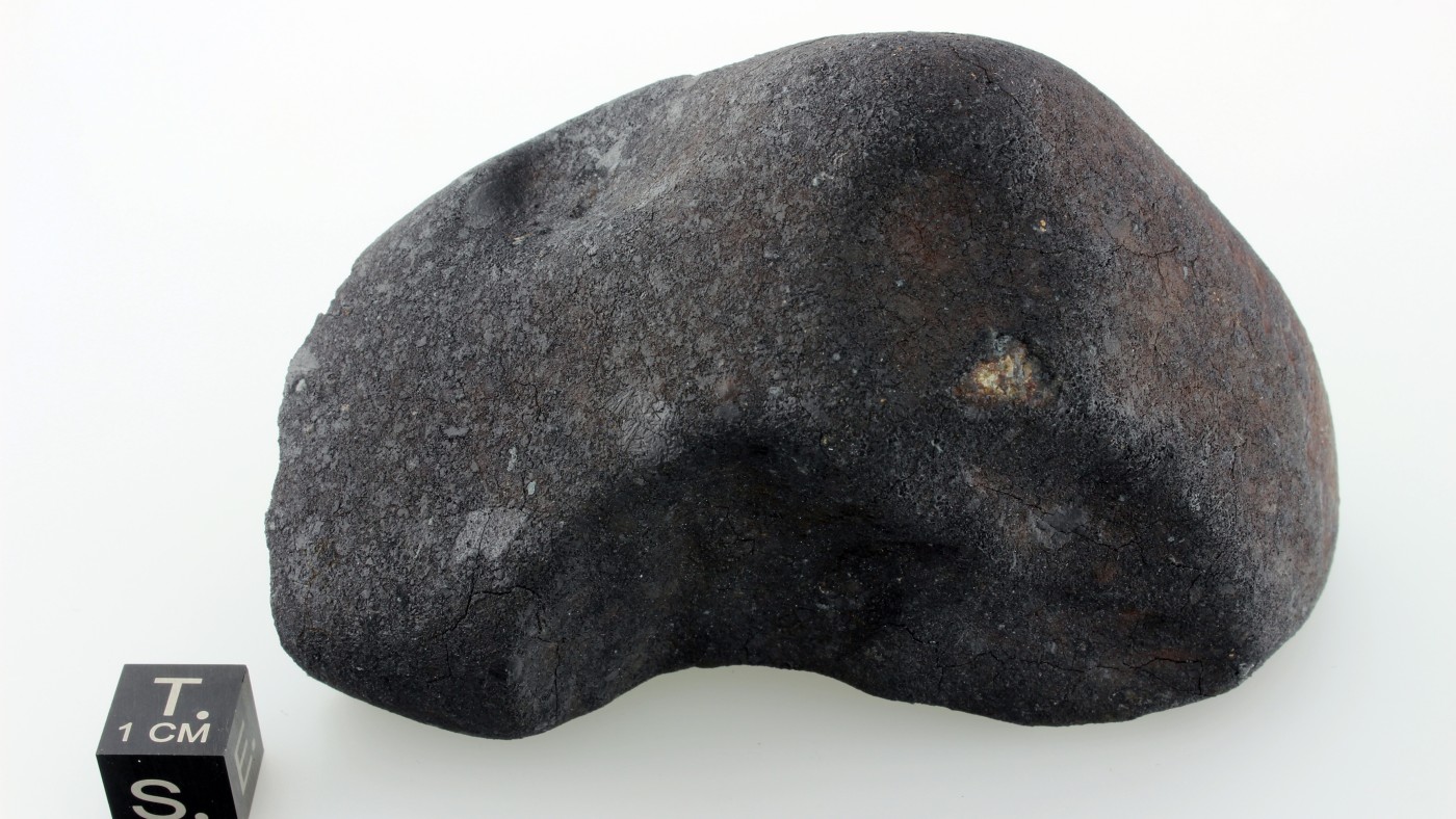 Dark, irregularly shaped meteorite with rough surface next to a black cube labeled 'T. 1 CM S.' (AI-generated alt-text,
                                 generated with GPT-4.1-mini)