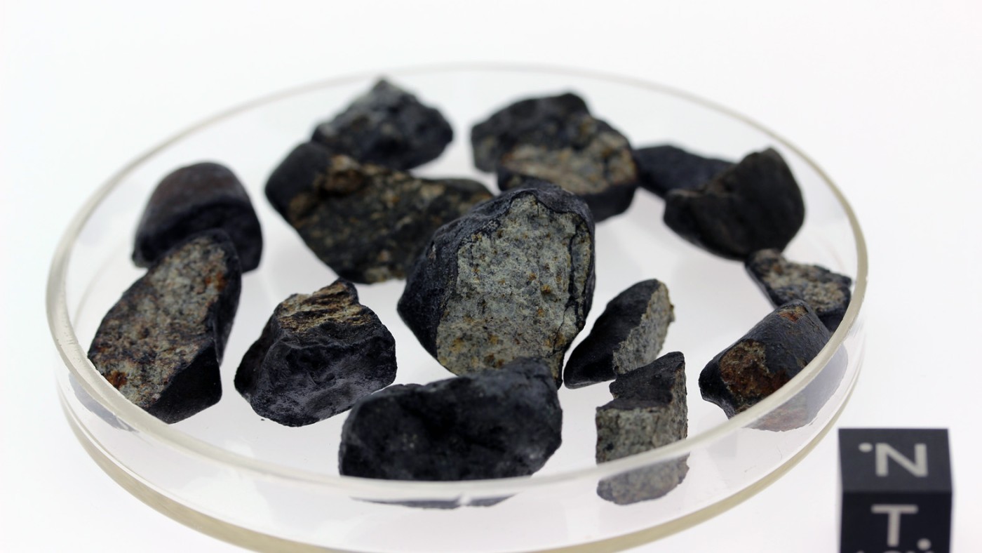 Several black and gray fragments of the Chelyabinsk meteorite in a shallow glass dish; scale cube marked 'N' and
                                 '1 CM' at lower right. (AI-generated alt-text, generated with GPT-4.1-mini)