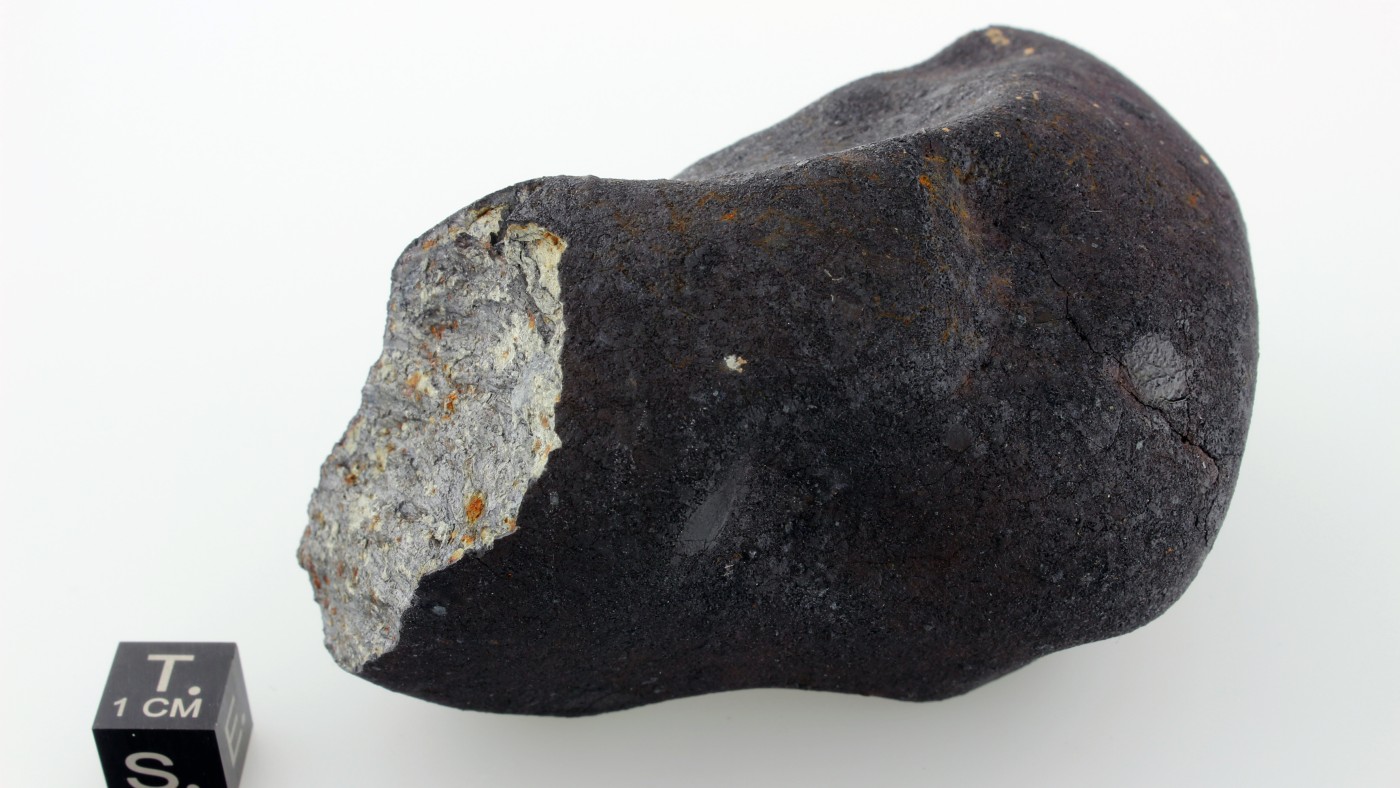 Dark, irregularly shaped meteorite with light fracture surface, cube with scale and letters beside it. (AI-generated
                                 alt-text, generated with GPT-4.1-mini)